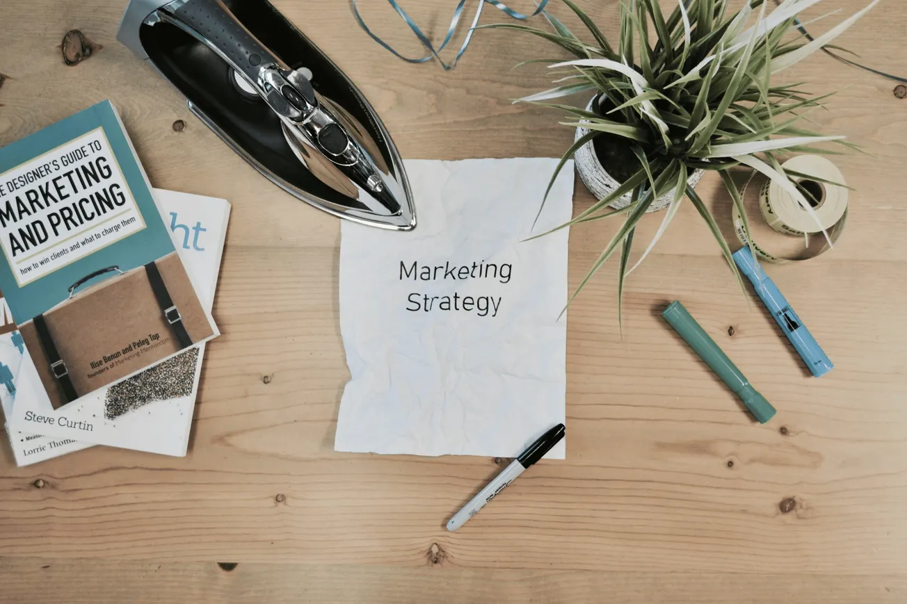 Marketing strategy planning on wooden desk with books, markers, iron, plant, and crumpled paper.
