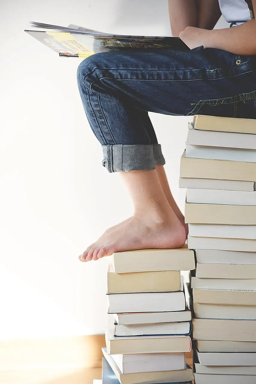 Stack of books with a person sitting on top, reading a magazine, casual jeans, and barefoot, representing reading, learning,