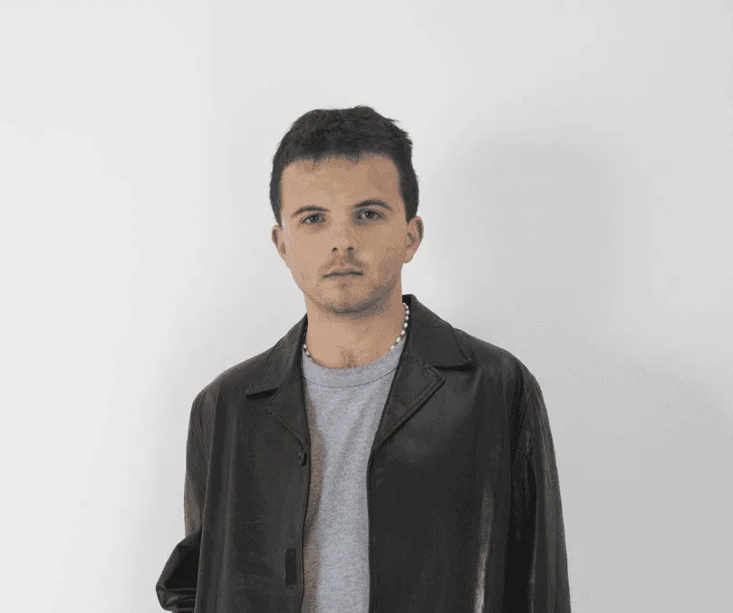 Fashion-forward young man wearing a black leather jacket and gray t-shirt, standing against a plain white background, represe