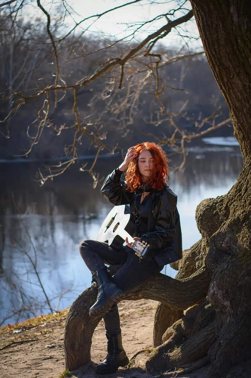 Moody young woman with red curly hair sitting on a tree branch by the lake, holding an acoustic guitar, wearing black boots a