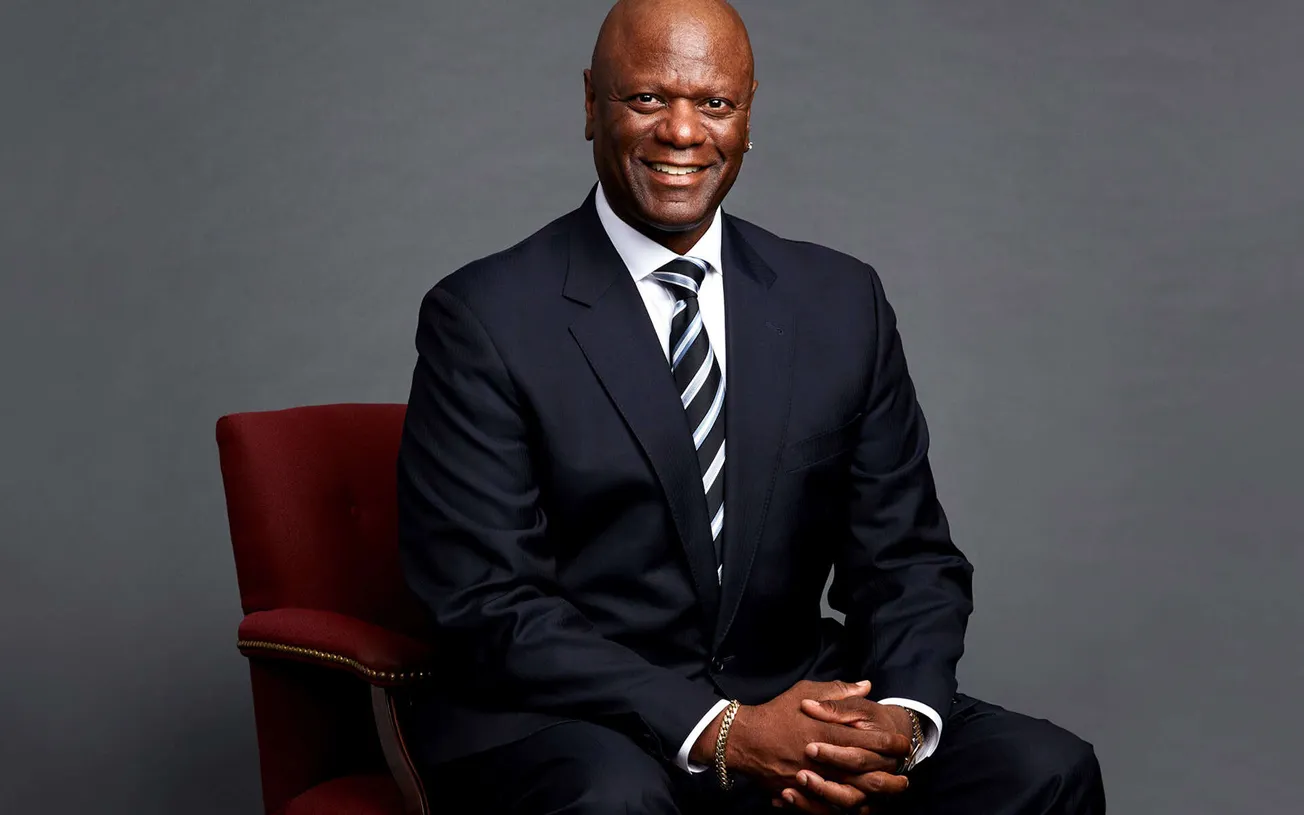 Senior African American man in formal suit sitting on a red chair against a neutral gray background, smiling confidently show