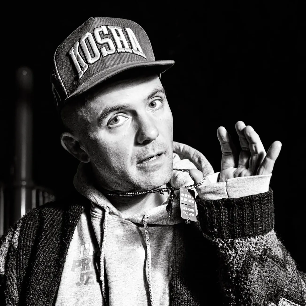 Young man wearing a "KOSH" cap, hoodie, and sweater, making a hand gesture, candid style, black and white photograph, express