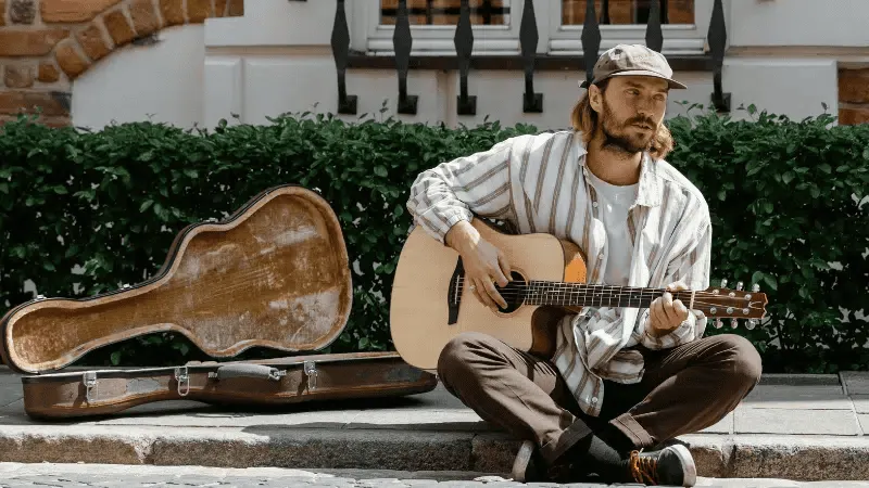 Street musician playing acoustic guitar on urban sidewalk, open guitar case for tips, casual outdoor scene, capturing street