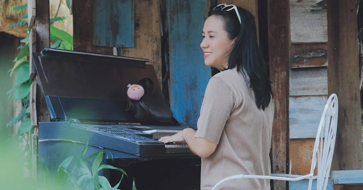 A woman smiling while playing an outdoor piano, surrounded by lush greenery and rustic wooden walls, emphasizing music, outdo
