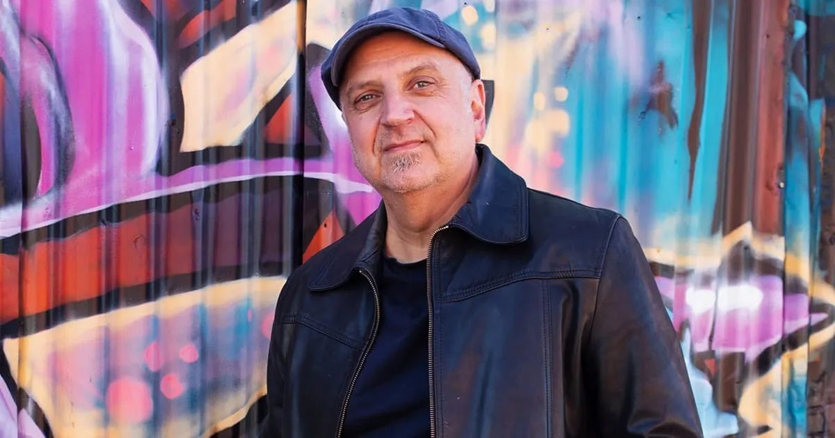 Older man in black leather jacket standing in front of colorful graffiti wall, urban street style, confident expression, musi
