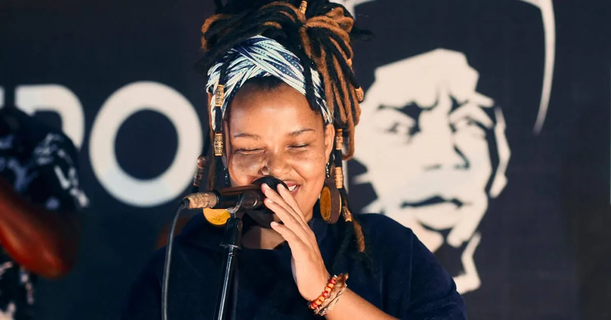 A vibrant female singer with dreadlocks and colorful headwrap performing passionately into a microphone at a music event.