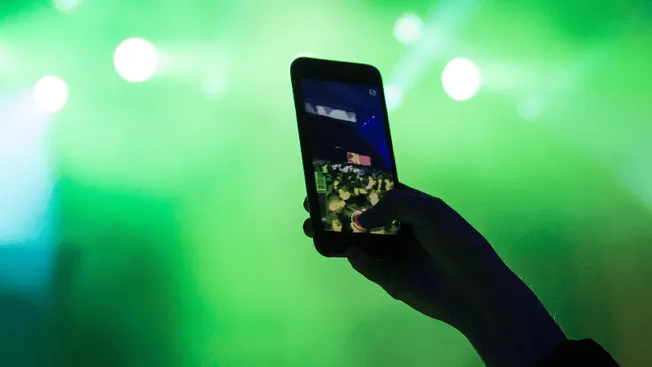 A person holding a smartphone capturing or streaming live music concert with bright green stage lighting in the background.