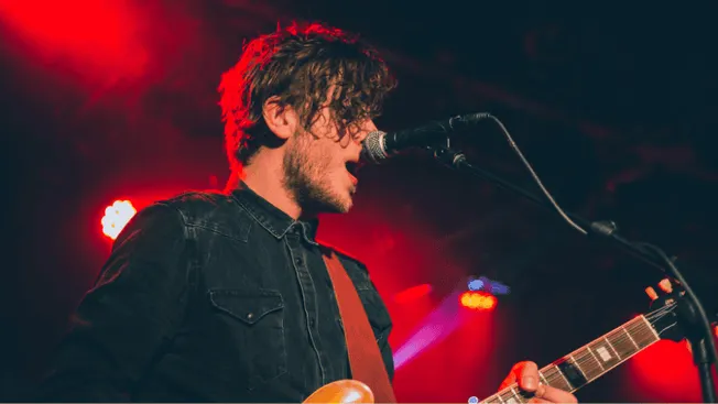 Soft-focus photo of a young male musician playing an electric guitar and singing into a microphone on stage with vibrant red