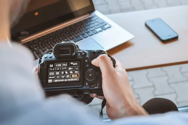 High-quality DSLR camera on photographer's hand with laptop and smartphone on wooden desk, focusing on digital media producti