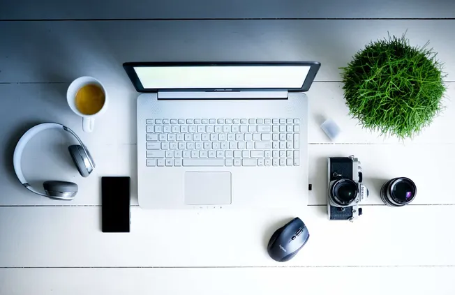Clean workspace with laptop, camera, headphones, smartphone, mouse, tea, and plant on white wooden desk.