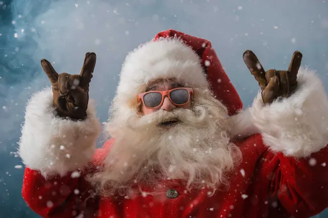 Santa Claus in a winter setting wearing sunglasses, making rock and roll hand gestures amidst falling snow, holiday fun, and