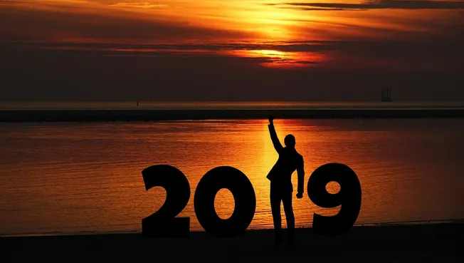 Silhouette of a person celebrating New Year 2019 at sunset on the beach with large 2019 numbers in the foreground, creating a