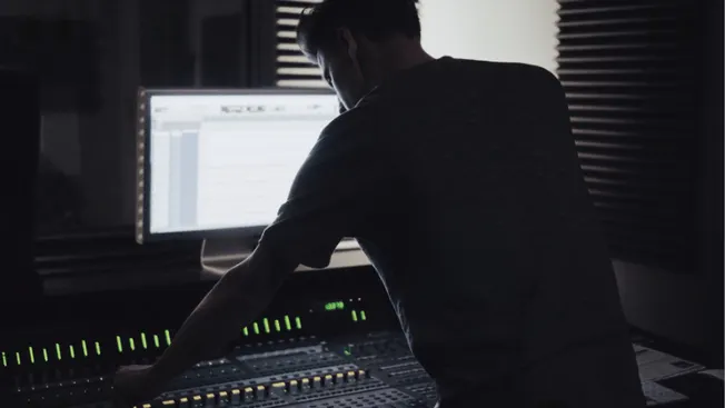 Professional music producer working at a mixing console with a computer in a dark studio environment.