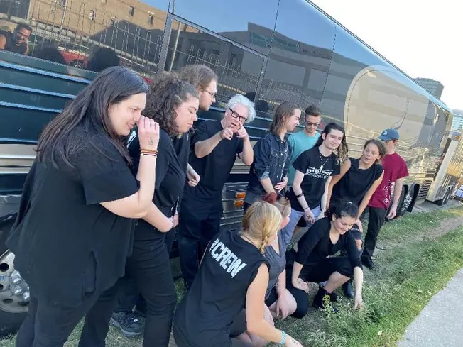 A diverse group of young people and an older woman gather outdoors beside a large black bus, engaging in a lively discussion