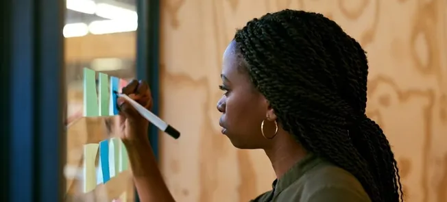 Post-it notes on glass wall with woman writing and planning during brainstorming session.