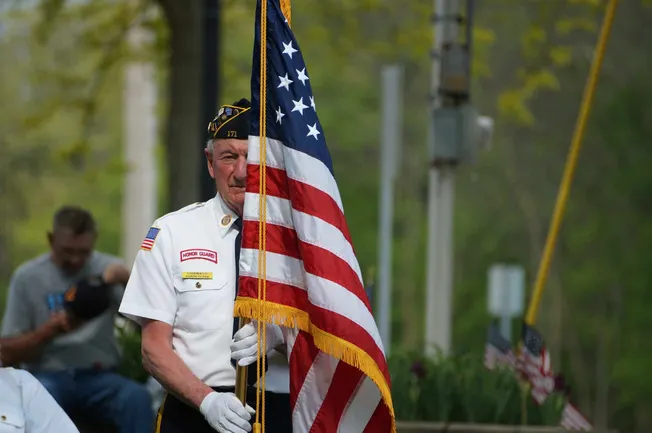 Veteran honor guard member holding the American flag during Memorial Day ceremony in a park with trees in the background.