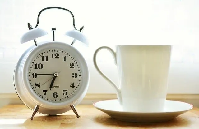 Vintage white alarm clock next to a white coffee cup and saucer on a wooden surface, morning breakfast setting, natural light