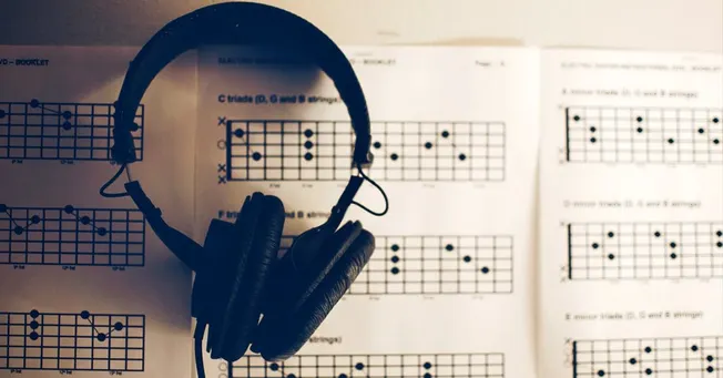 Guitar fretboard charts and a black over-ear headphone listening to music on sheet music background for musicians.