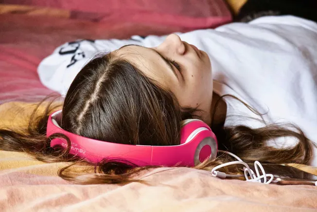Close-up of a young woman sleeping on a bed with pink headphones, resting peacefully with eyes closed and brown hair spread o