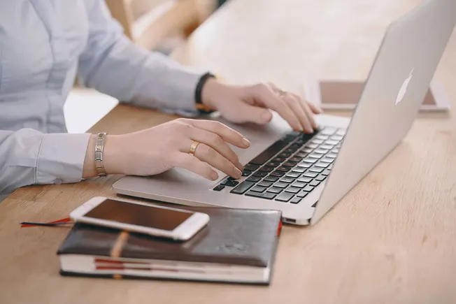 Professional woman working on a laptop at a wooden desk with a smartphone and notebooks, emphasizing productivity and digital