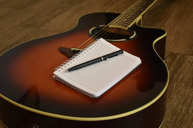 A vintage acoustic guitar with a lined notepad and black pen resting on it, placed on a wooden floor, ideal for music songwri