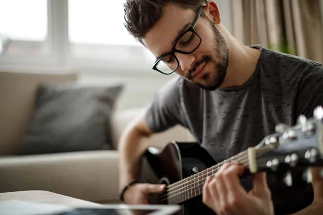 Young man playing acoustic guitar at home, practicing music, relaxing, inner creativity, comfortable living room, musical hob