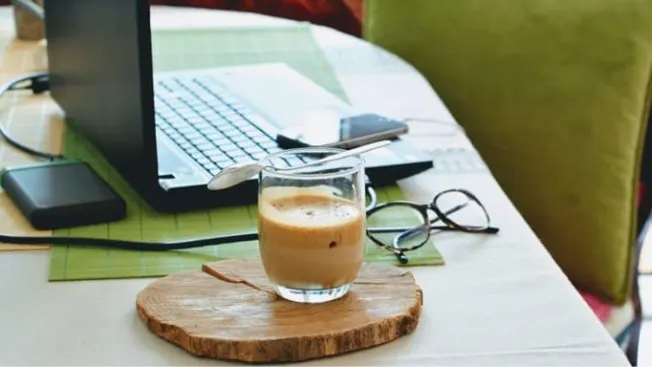 A steaming glass of coffee on a wooden coaster placed on a desk with a laptop, glasses, and a smartphone, capturing a cozy wo