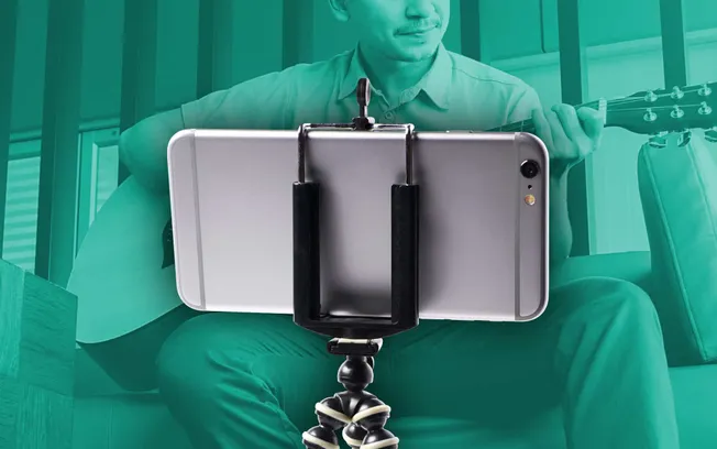 Close-up of a person playing an acoustic guitar while recording a video on a smartphone mounted on a flexible tripod. The sce