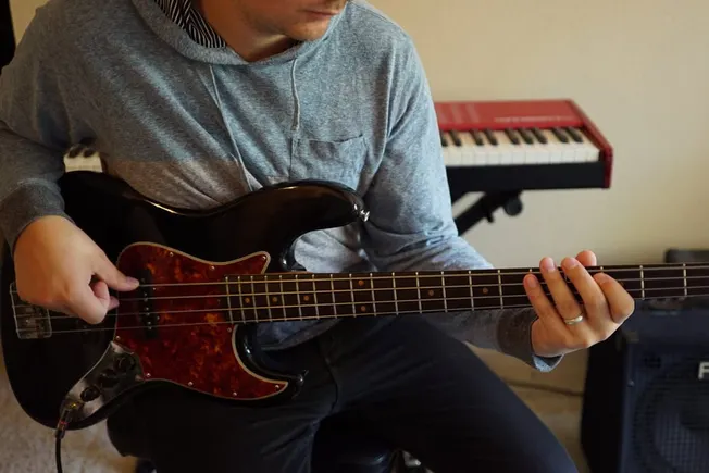 Electric bass guitar playing musician in home music studio with keyboard and amplifier in background.
