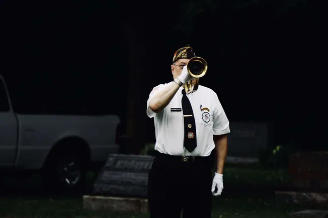 Patriot playing bugle at a memorial ceremony honoring military veterans, wearing ceremonial uniform with badges and patriotic