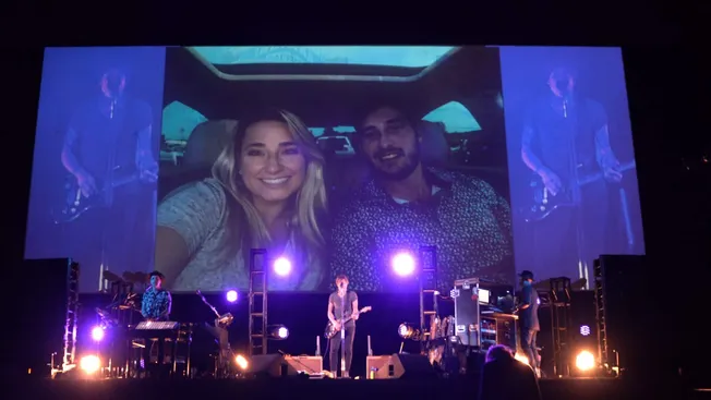 Live concert with band performing on stage, large screen displaying audience photo, colorful stage lighting creating vibrant
