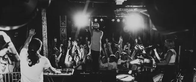 Vibrant live concert scene with a DJ, keyboardist, and enthusiastic audience raising hands at an intimate music venue. Black