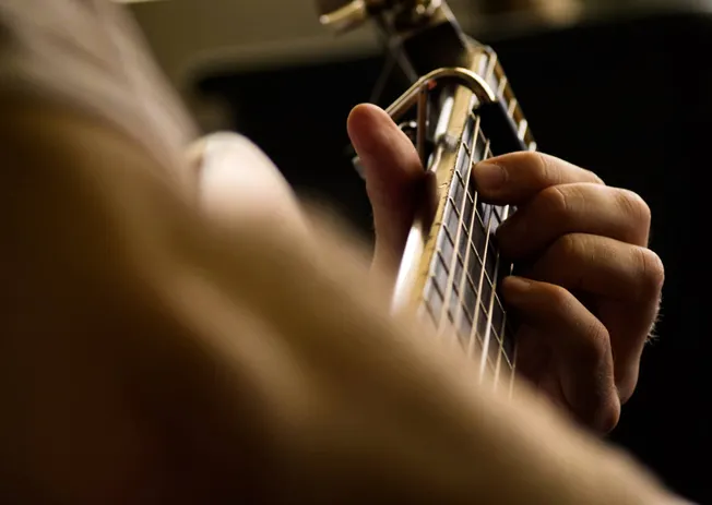 A person playing an acoustic guitar, focusing on their hand on the fretboard, captured in warm lighting. The image highlights