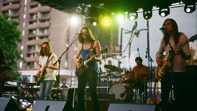 Bright outdoor concert featuring a live band performing on stage with guitars and drums during a festival.