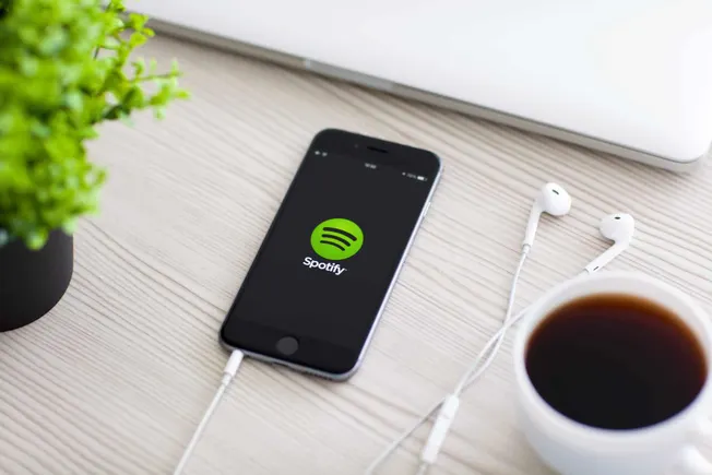 Spotify app on smartphone with earphones, coffee, and laptop on a light wooden desk, representing music streaming and digital