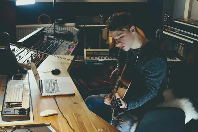 Young male musician playing acoustic guitar in a professional recording studio with musical equipment and keyboards.