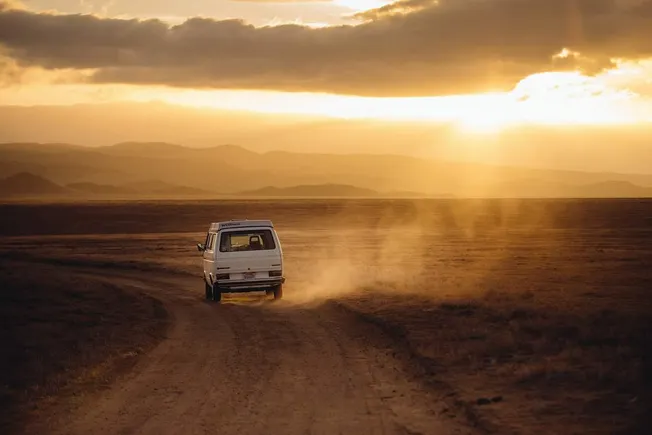 A vintage white van driving on a dusty dirt road through an open desert landscape during sunset with mountains in the backgro