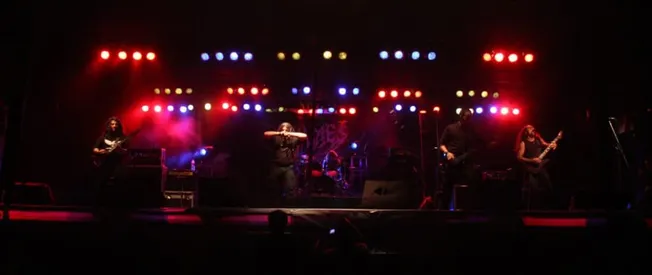 Guitarists and drummer performing live on stage with vibrant red, blue, and yellow stage lighting, showcasing a dynamic conce