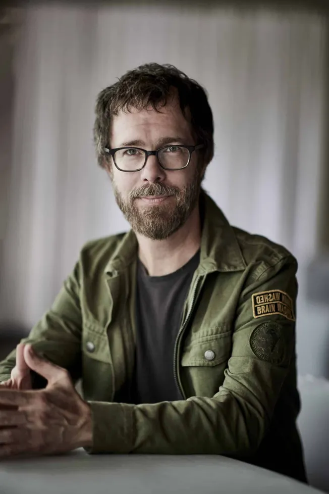 Thoughtful man with glasses and beard wearing a military-style jacket, sitting indoors, representing music industry, creative