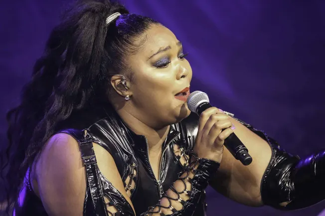Vocalist performing live on stage, wearing a black leather outfit, with bold makeup and long wavy hair, holding a microphone,