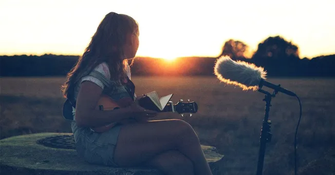 A young woman playing guitar outdoors at sunset, recording vocals with a professional microphone, creating music in a natural