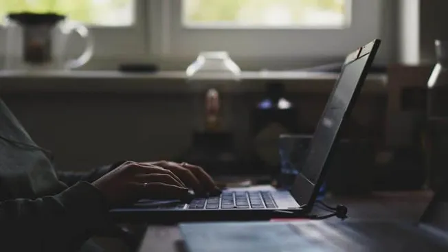 Person typing on a laptop in a dimly lit workspace, with natural light coming from the window. Focus on digital work, technol