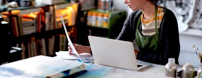 Young woman working on a creative project with a laptop and printed papers in a cozy, modern workspace.