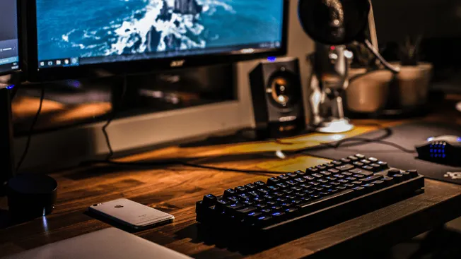 A modern desktop setup featuring a computer monitor, mechanical keyboard, smartphone, and audio equipment on a wooden desk in