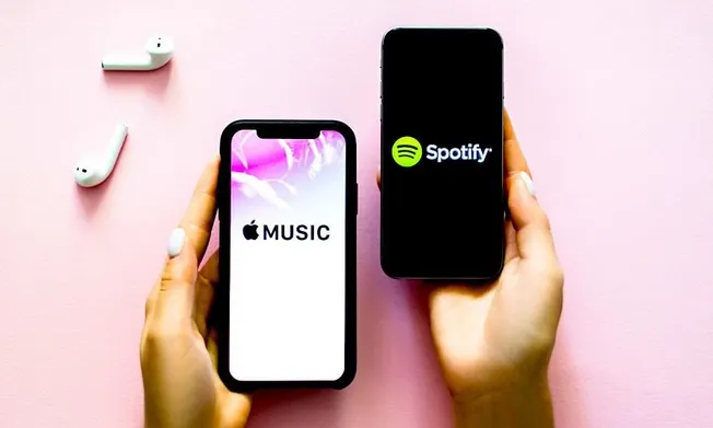 Apple Music and Spotify app screens displayed on smartphones with wireless earbuds on a pink background, highlighting popular
