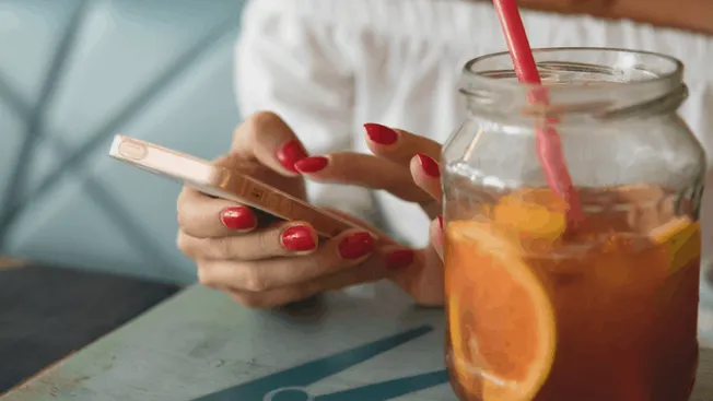 She's using her smartphone with a drink containing orange slices on a wooden table at a cafe.