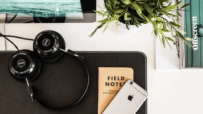 Headphones, a smartphone, a field note, and a plant on a workspace desk, capturing a modern music and content creation setup