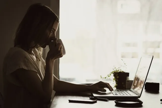 Cozy woman working on laptop in natural light at home, sipping coffee, freelance, remote work, modern workspace, productivity