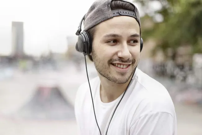 Modern young man smiling while wearing headphones outdoors, enjoying music, urban background, casual style, lifestyle, leisur