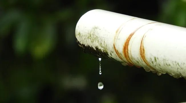 Fallen rainwater drip from a weathered white PVC pipe with rust stains, close-up shot, natural background, emphasizing water